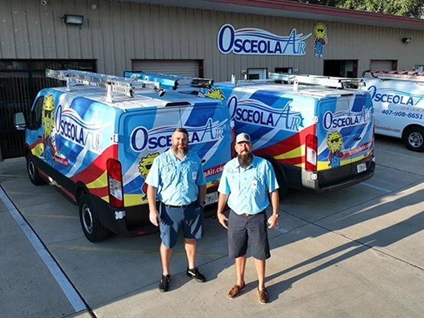Two men in blue uniforms stand in front of Osceola Air vans parked outside a commercial building with Osceola Air signage.