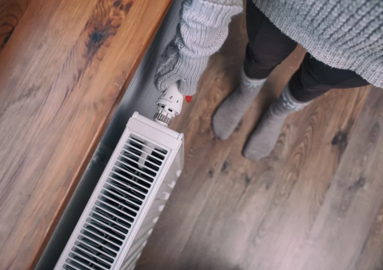 Person in warm clothing adjusts the thermostat on a white radiator, standing on a wooden floor next to a wooden surface.