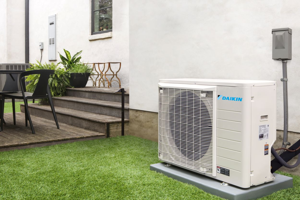 Daikin outdoor mini-split HVAC unit installed on a concrete pad next to a modern home with white exterior walls, surrounded by artificial turf, potted plants, and a small wooden deck with outdoor seating.