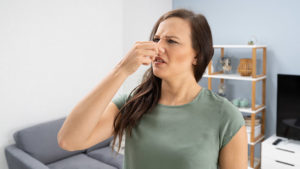 Woman standing in a living room, pinching her nose and looking uncomfortable, suggesting she smells something unpleasant.