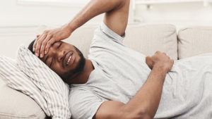 A man lying on a couch with his eyes closed, holding his forehead and appearing to be in pain or discomfort.