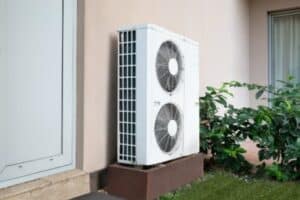 A white outdoor air conditioning unit with two fans is mounted on a stand next to a building, near a door and some green plants.