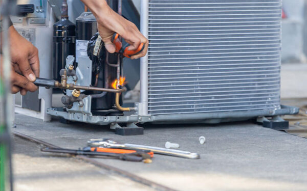 A person uses a torch to weld pipes on an air conditioning unit, with various tools lying on the ground nearby.