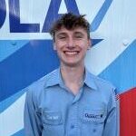 A young man in a light blue uniform shirt stands smiling in front of a wall with blue, red, and white geometric designs.
