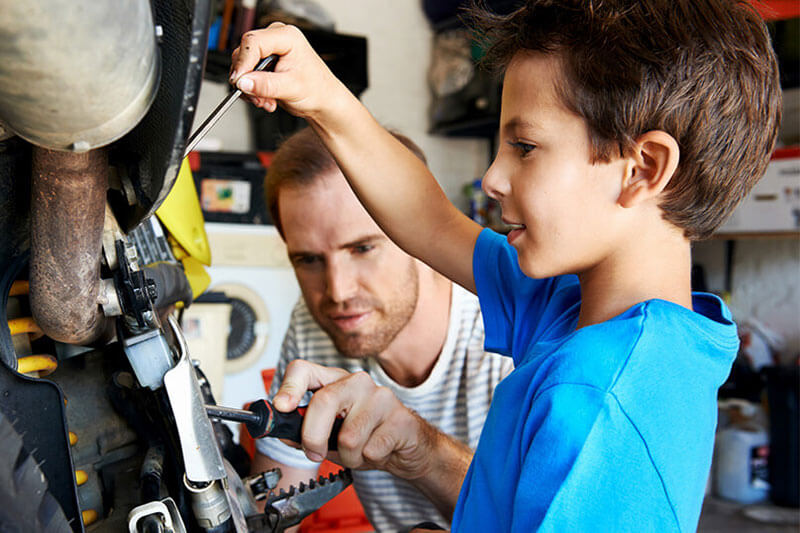 A man and a boy use screwdrivers to work on a motorcycle together in a garage.