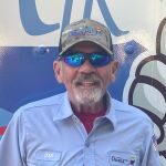 An older man with a gray beard wearing a cap, blue reflective sunglasses, and a light blue shirt stands in front of a colorful background with graphics.