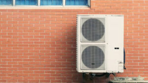 An outdoor air conditioning unit is mounted on a red brick wall beneath a window with blue glass panes.