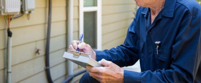 A person in a blue uniform writes on a clipboard while inspecting equipment outside a building.