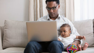 A man sits on a couch working on a laptop with a baby on his lap holding a colorful toy.