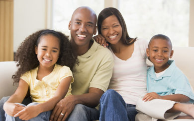 A family of four sits closely together on a couch, smiling at the camera in a well-lit living room.