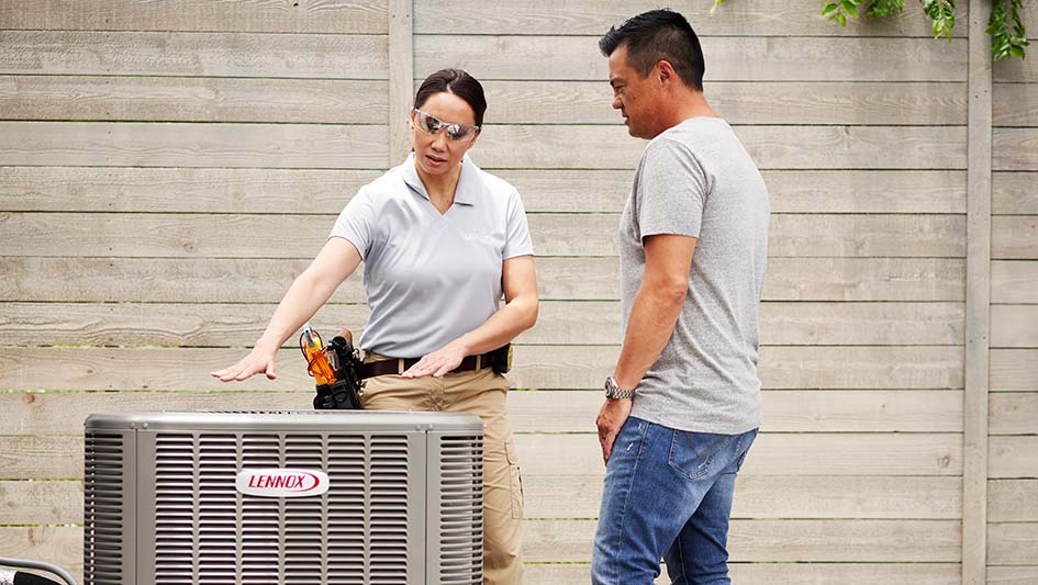 A technician explains an outdoor Lennox air conditioning unit to a man, standing next to a wooden fence.