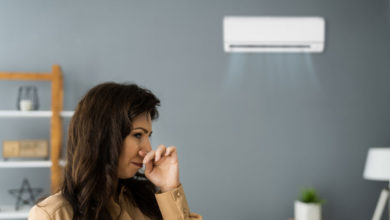 Woman indoors pinching her nose, standing near an air conditioner mounted on the wall, with cool air flowing from the unit.