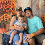 Meet Our Team: A woman in a cap and overalls, a man in a light blue shirt, and their baby in striped clothes sit together in front of a sunflower backdrop.