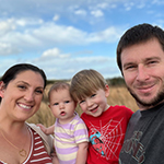 Meet Our Team: A woman, a man, and two young children pose together outdoors in front of tall grass under a partly cloudy sky.