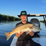 Meet our team: A person wearing a black hat and sunglasses sits on a boat, holding a large fish with both hands, with water and trees in the background.