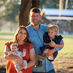 Meet our team: A man and woman stand outdoors, smiling at the camera, each holding a young child. Trees and a structure are visible in the background.