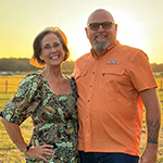 Meet Our Team: A smiling man and woman stand together outdoors in a field at sunset. The woman wears a patterned dress and the man wears sunglasses and an orange shirt.
