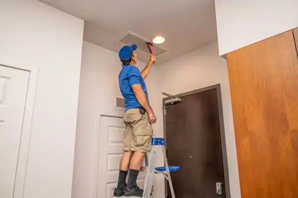 A person standing on a stepladder cleans an air vent on the ceiling in a residential hallway.