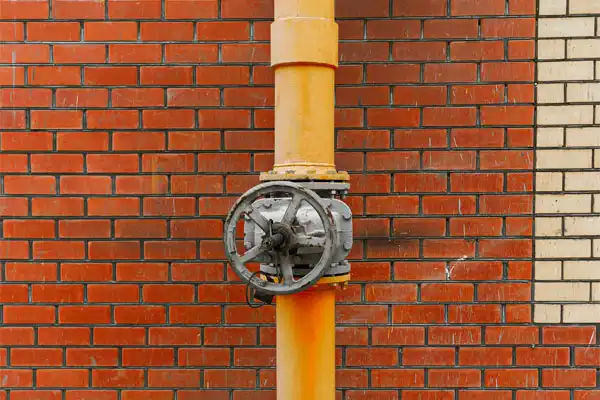A yellow industrial pipe with a metal valve wheel is mounted on a red and beige brick wall.