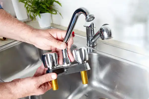 A person holds a chrome kitchen faucet fixture with two handles above a stainless steel sink.