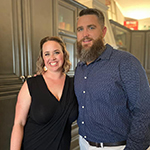 Meet Our Team: A woman in a black dress and a man in a blue shirt stand side by side, smiling, in a kitchen with dark cabinets.