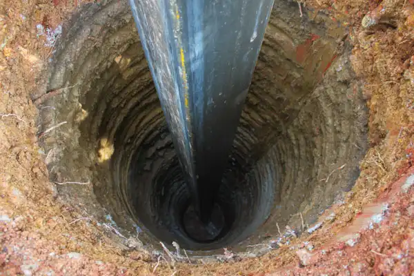 A large pipe being inserted vertically into a freshly dug, circular hole in the ground, surrounded by exposed soil layers.