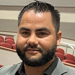 Meet Our Team: A man with short dark hair and a beard, dressed in a gray blazer, stands indoors in front of red auditorium seats.