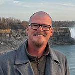 Meet Our Team: A man with glasses and a goatee stands outdoors near a waterfall and rocky shoreline, wearing a grey coat and smiling at the camera.
