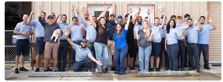 A group of about 20 people, some in uniforms, pose and wave outside a beige industrial building; one person holds a small dog.