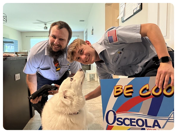 Two uniformed workers pose indoors with a white dog; one holds a sign reading "BE COOL" and "Osceola" with a colorful design.