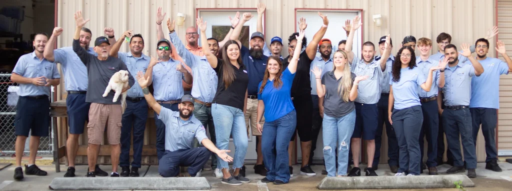 A group of twenty-one people, mostly in work uniforms, and one small dog, pose outside a building, smiling and raising their hands.