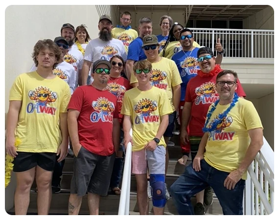 A group of people pose on stairs, wearing matching yellow and red "Oh Way" t-shirts and sunglasses, some with leis and hats, in an outdoor setting.