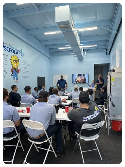 A group of people in uniform shirts sit at tables in a blue-walled room, listening to a presentation with slides displayed on a TV screen at the front.