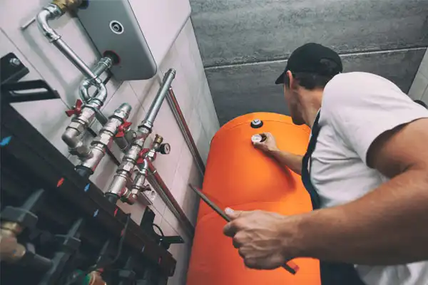 A worker in a cap adjusts a valve on a large orange water heater tank in a utility room with visible pipes and gauges.