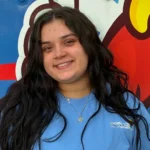 Meet Our Team: A woman with long dark hair wearing a blue Osceola-branded shirt smiles at the camera, standing in front of a colorful abstract background.