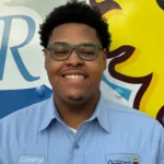 Meet Our Team: A person wearing glasses and a light blue work shirt with a name tag reading "Cameron" stands smiling in front of a colorful mural.