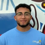 Meet Our Team: A man wearing glasses and a light blue "Osceola" shirt stands in front of a colorful mural, looking at the camera and smiling slightly.