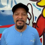 Meet Our Team: A man with a goatee and mustache, wearing a blue T-shirt and black cap, stands in front of a colorful mural with a pen clipped to his shirt.