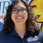 Meet Our Team: A woman with curly hair and glasses, smiling, wearing a navy blue Osceola Air AC & Plumbing Pros polo shirt, stands in front of a colorful company vehicle.