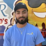 Meet Our Team: A man with a beard, wearing a blue Osceola t-shirt, black cap, and gold cross necklace, stands in front of a colorful ice cream truck graphic.